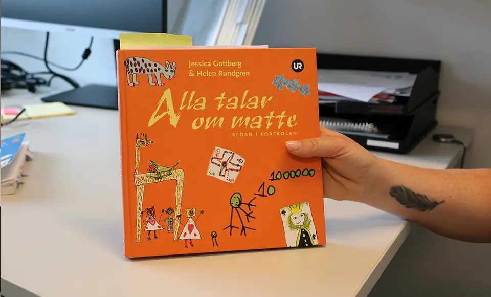 En hand håller upp boken Alla talar om matte redan i förskolan på ett skrivbord.