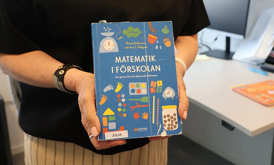 En person håller upp boken Matematik i förskolan framför ett skrivbord.