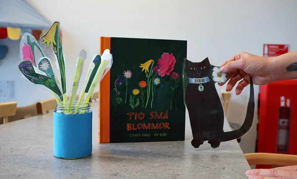 En hand håller en pappersfigur av en katt intill boken Tio små blommor och utklippta blommor i en burk.