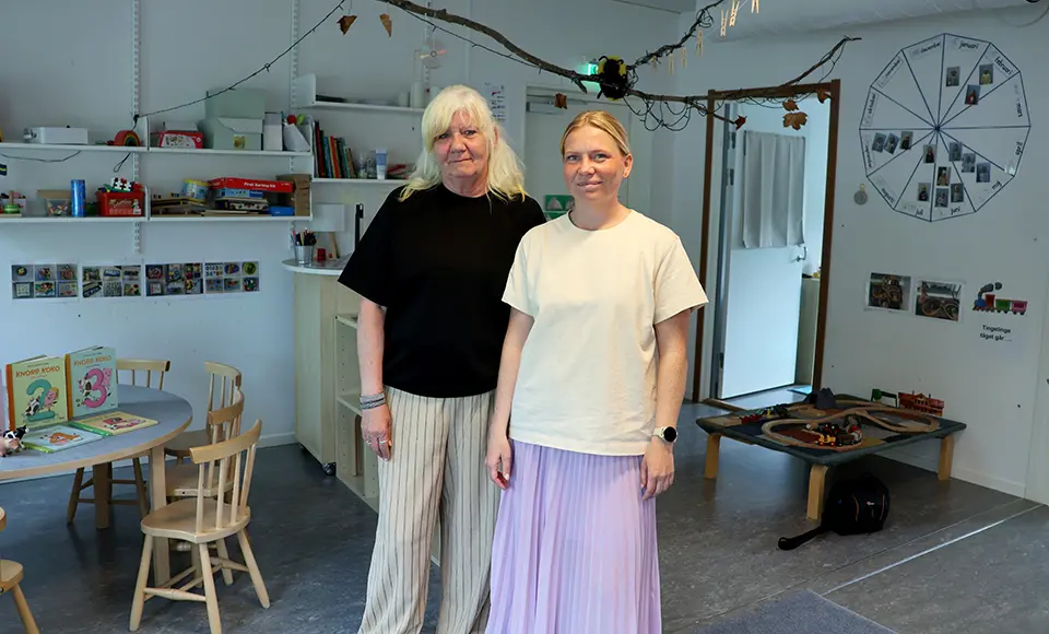 Två personer står i en förskolemiljö med matematikmaterial, barnböcker och bildstöd i bakgrunden.