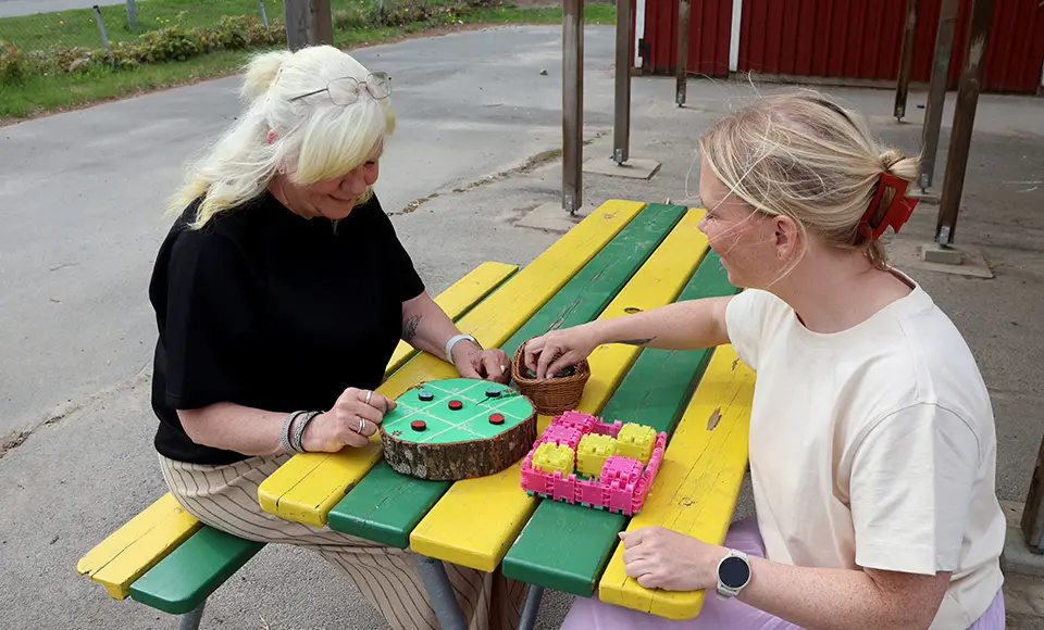 Två personer sitter vid ett bord utomhus med spelmaterial på en förskolegård.