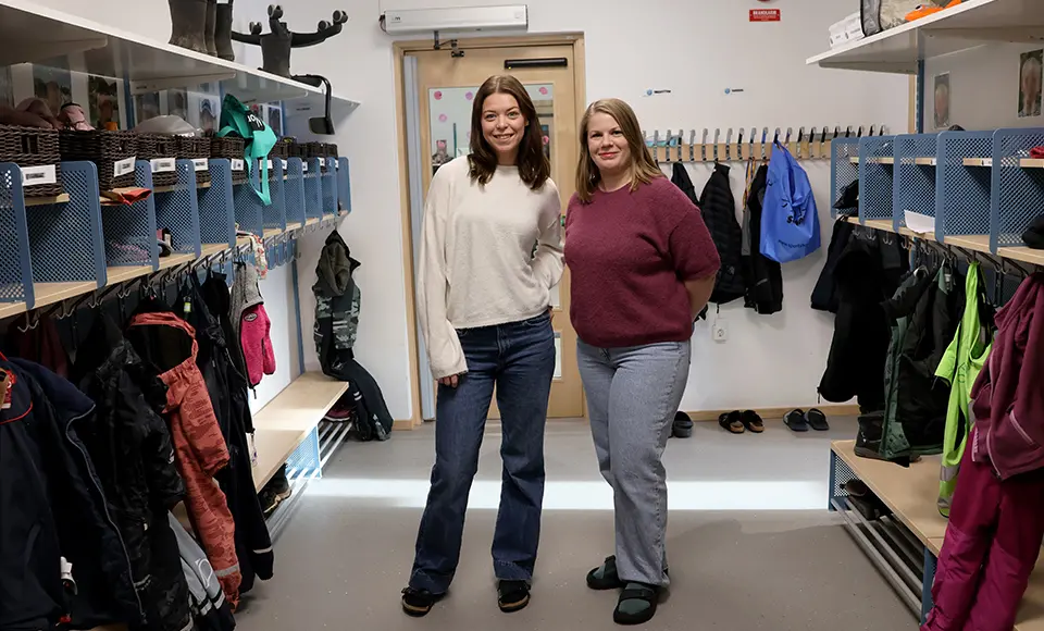 Emelie och Maria står bredvid varandra i en hall på förskolan. På hyllor och krokar hänger barnens ytterkläder.