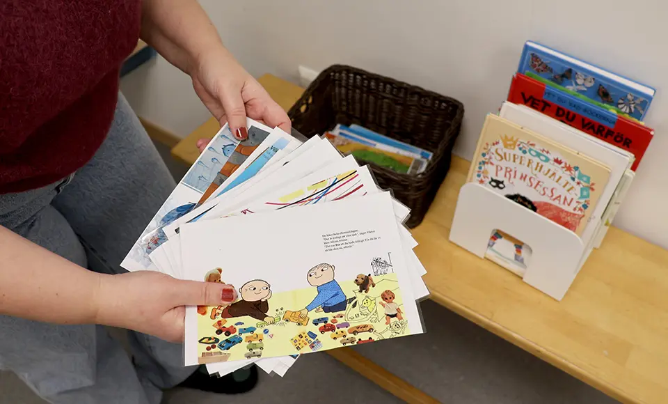 En pedagog håller i en bunt kort med bilder och korta texter. På en bänk bredvid står en korg med fler kort samt ett ställ med barnböcker, avsedda som material för barn under väntetiden i hallen.