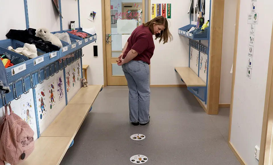 En pedagog står i en förskolehall mellan två långa bänkar med klädfack ovanför. På golvet framför pedagogen finns runda markeringar som visar var barn kan stå och vänta. Hallen är ordnad och anpassad för på- och avklädning.