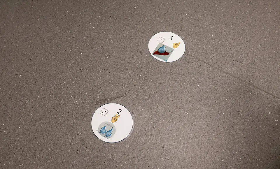 Två runda golvmarkeringar på ett grått golv visar turordning med siffror och illustrationer av figurer från boken Droppen Dripp. Markeringarna används som stöd för barn att veta var de kan stå och vänta.