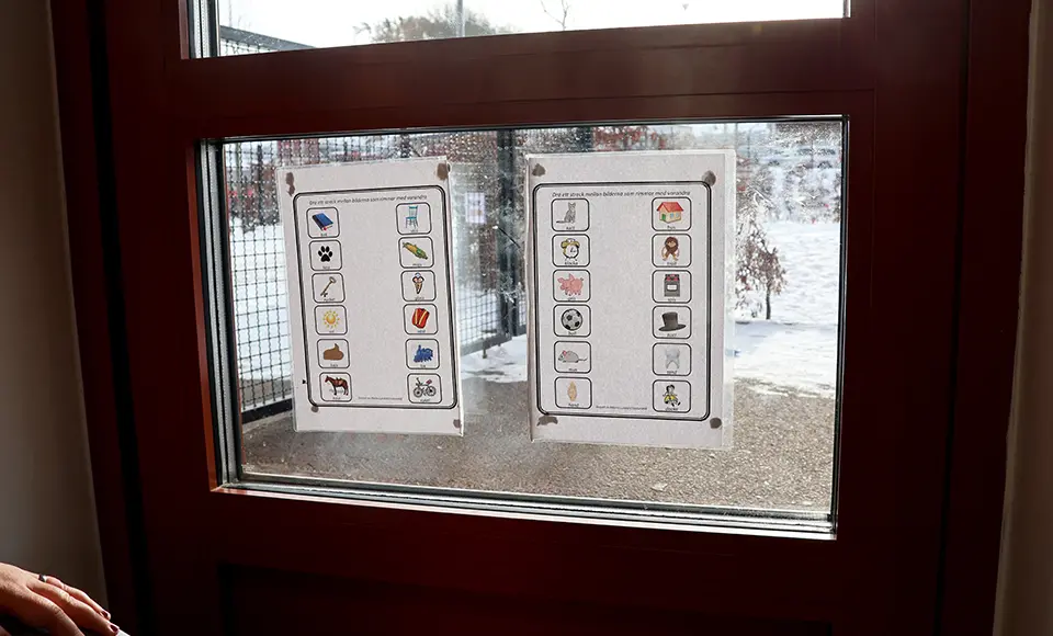 Två laminerade blad med rim och bildsymboler sitter uppsatta på ett fönster i en dörr. Genom fönstret syns en snötäckt gård utanför.