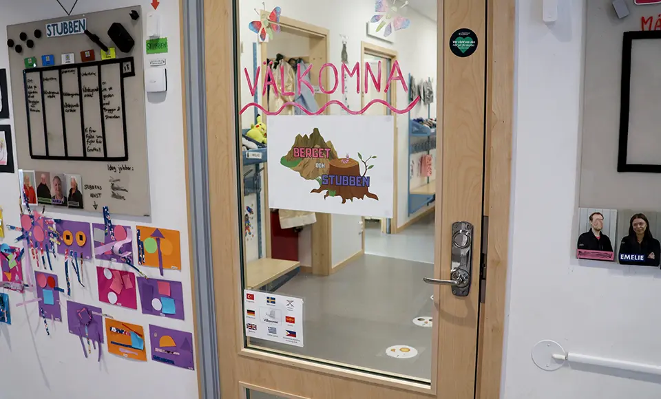 En glasdörr in till en förskoleavdelning med texten ”Välkomna” och skylten ”Berget och Stubben” på glaset. Genom dörren syns hallen med bänkar, klädfack och golvmarkeringar som visar var barn kan stå. Runt dörren finns väggar med barnens bilder, veckoplanering och annan information.