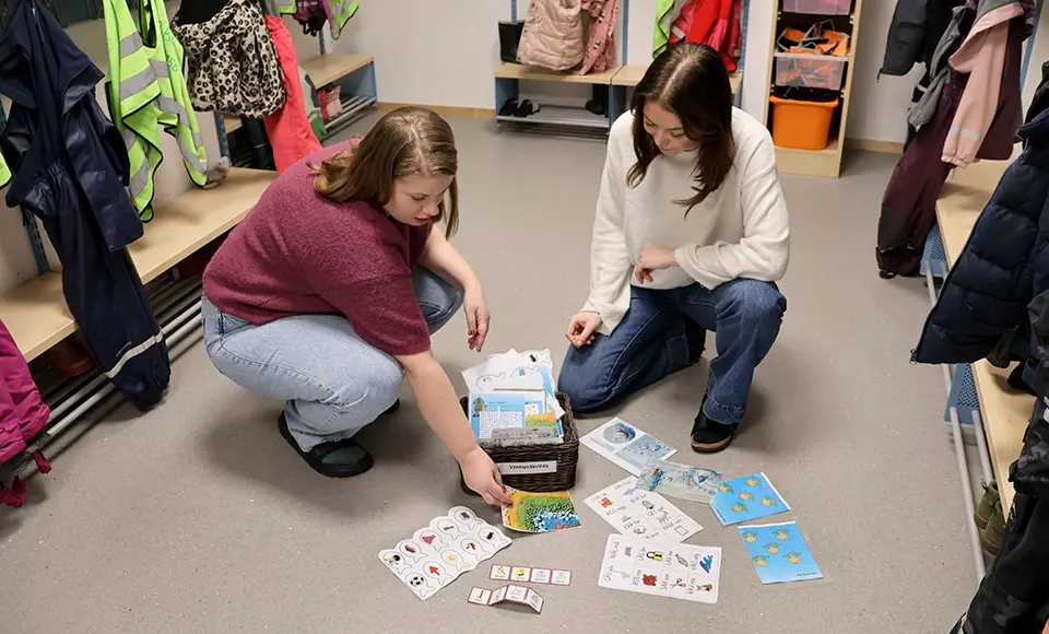 Två pedagoger sitter på golvet i en förskolehall med bänkar och ytterkläder längs väggarna. Framför dem ligger en korg med språkligt material, bildkort och böcker som är utspridda på golvet. Materialet används i hallen för att ge barnen något att titta på, samtala om och utforska under väntestunder.