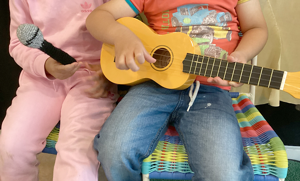 Två barn som sitter ner. Den ena har en mikrofon i handen, det andra barnet håller i en ukelele.