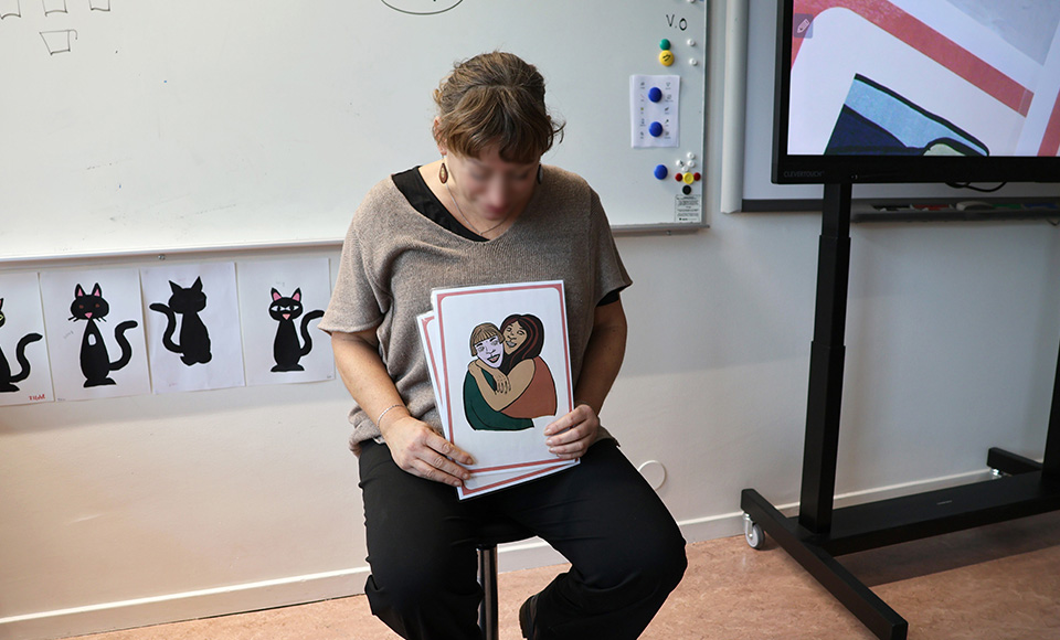 En person sitter längst fram i ett klassrum och håller i ett bildstöd som visar ett par som håller om varandra.