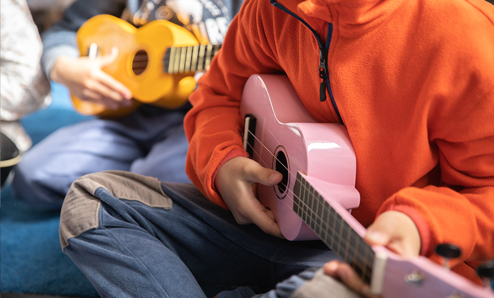 På bilden ser man överkroppen på barn som håller i varsin ukulele. De har färgglada tröjor på sig.