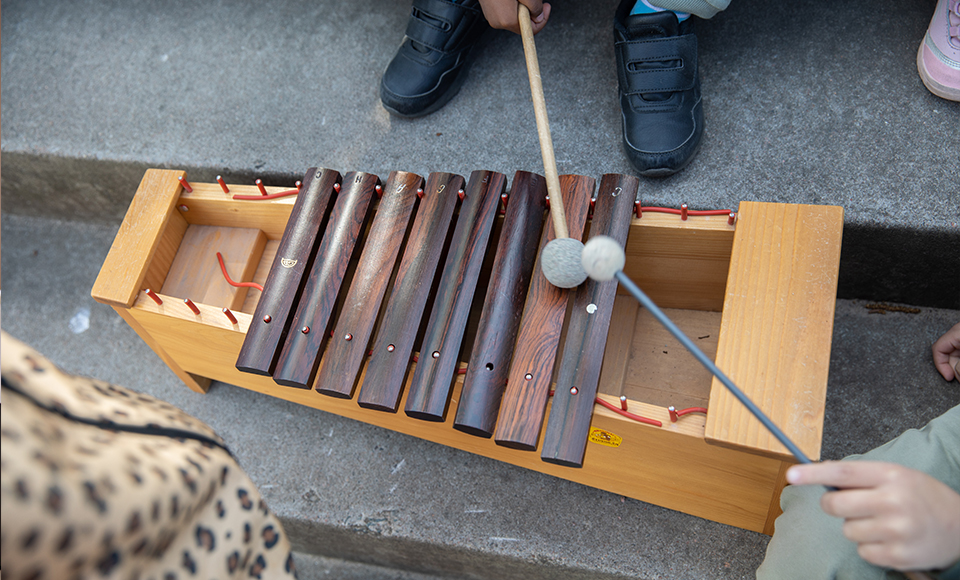 Foto på en xylofon tagen uppifrån. På bilden syns fötterna på barn som står runt omkring beredda på att spela.