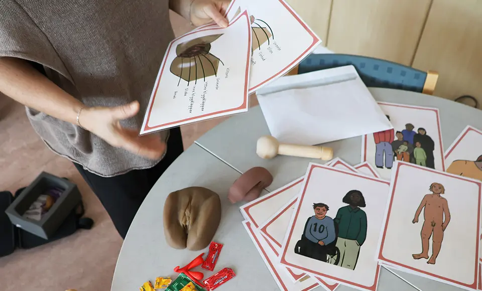 På ett bord ligger konkret material som verklighetstrogna modeller av kön, kondomer och bildstöd. Någon håller upp två bildstöd.
