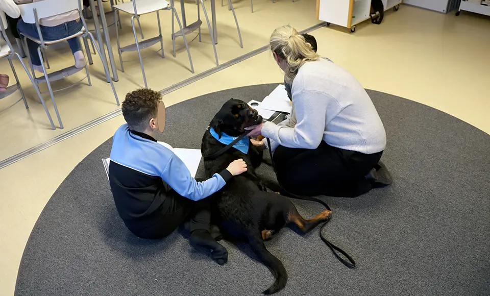 Två barn sitter på en matta. Emellan dem ligger hunden Börje och läraren sitter bredvid.