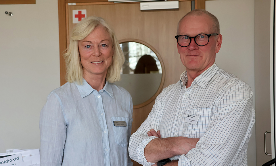 Två personer står bredvid varandra inomhus, framför en dörr med runt fönster och tittar in i kameran.