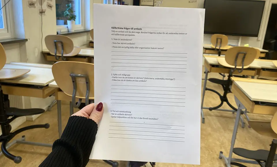 En hand håller upp ett arbetsblad med rubriken ’Källkritiska frågor till artikeln’ i ett klassrum med tomma stolar och bord i bakgrunden. Arbetsbladet innehåller frågor om vem som är avsändare, syfte och sammanhang för att analysera en text källkritiskt