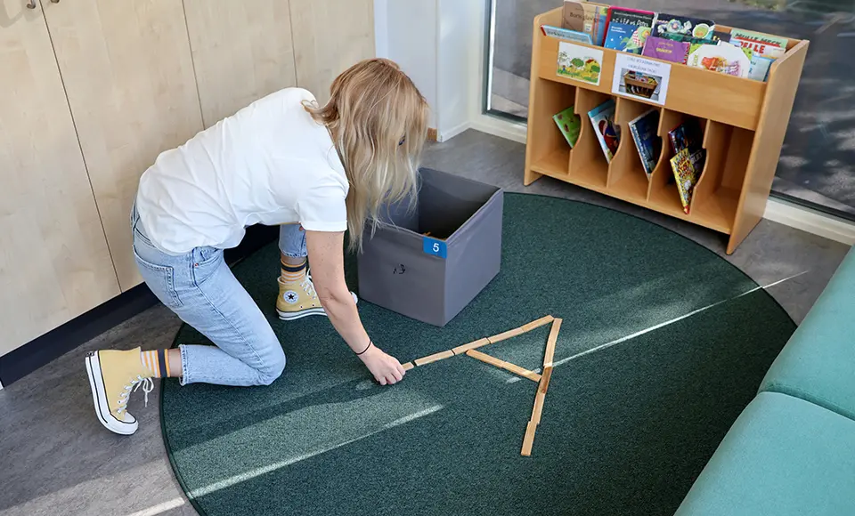 Malin sitter på huk på golvet och bygger bokstaven A av Kapplastavar.