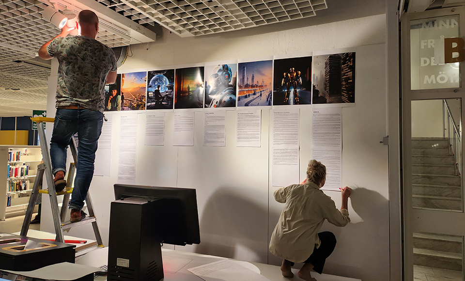 Två personer arbetar med att sätta upp en utställning på en vit vägg inomhus. En person står på en stege och justerar en röd lampa i taket, medan den andra sitter på golvet och fäster textark på väggen. Ovanför textarken sitter en rad färgstarka fotografier som visar futuristiska stadsmiljöer och teknikmotiv. I förgrunden syns en datorskärm på ett bord, och i bakgrunden finns bokhyllor med böcker.