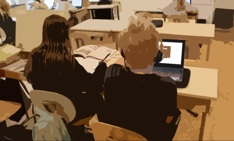 Två elever sitter bredvid varandra. Den ena skriver i en arbetsbok och den andra tittar på en film i datorn.