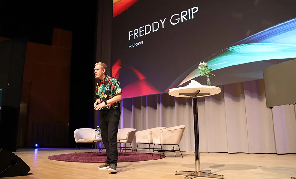 Freddy Grip går på scen. Han har en röd fluga och mönstrad skjorta i olika färger på sig.