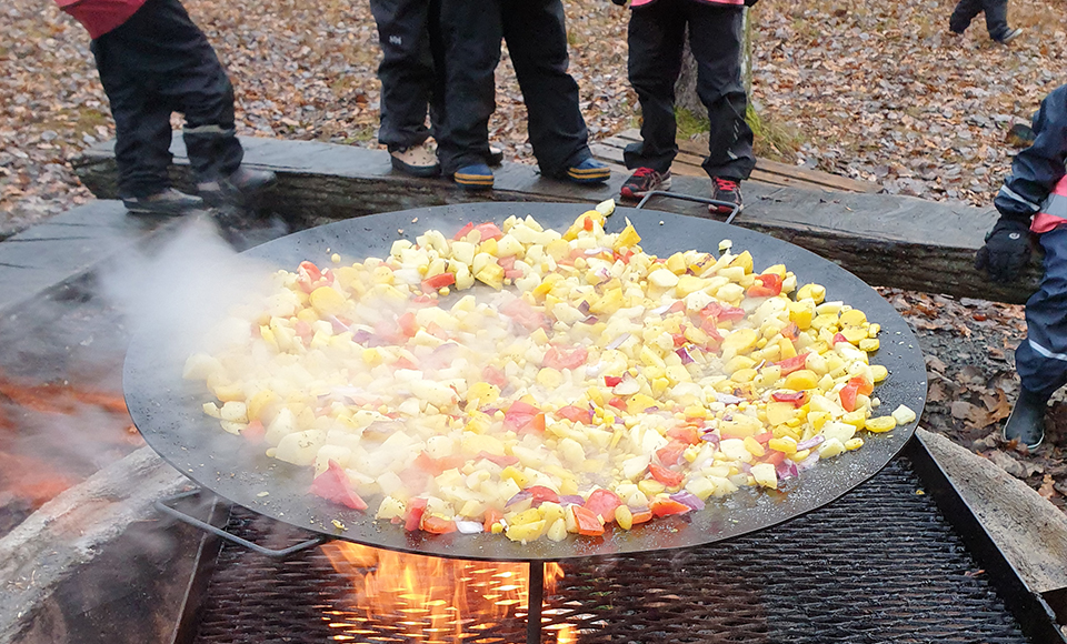 En wokpanna full med mat som står på en öppen eld. Det syns fötter från barn som står omkring.