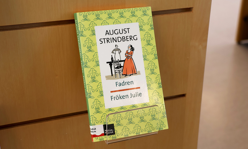 En bok med titeln "Fröken Julie" sitter uppställd med framsidan synlig i ett vägghängt bokställ av trä.