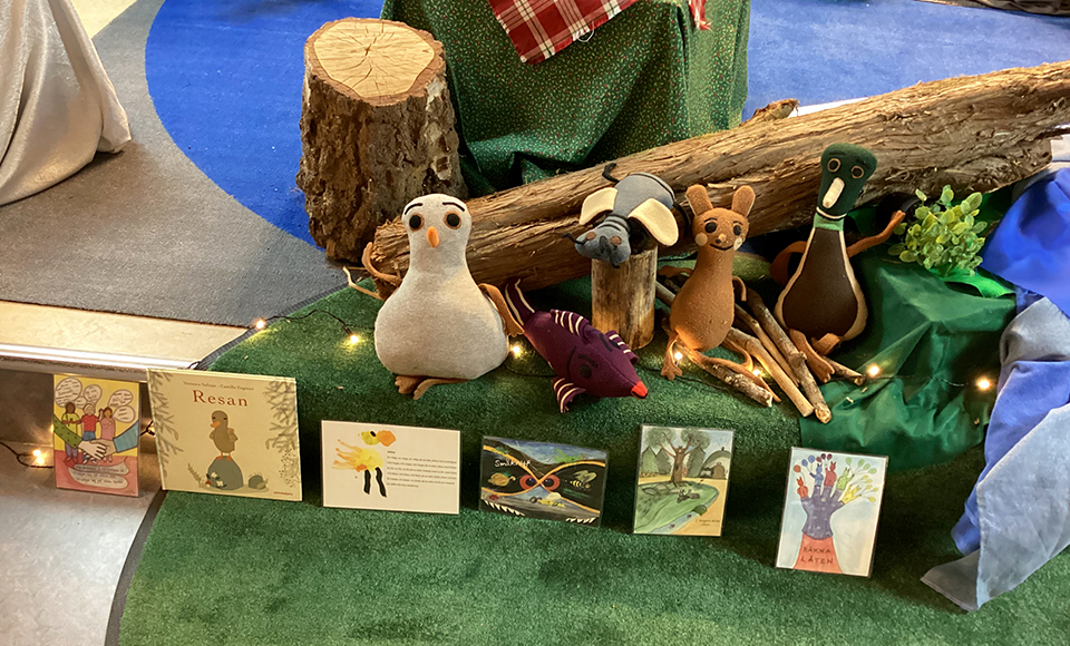 Ett arrangemang med mjuka djurfigurer framför en trädstam, omgivna av grön dekor och små ljusslingor. Framför figurerna står flera färgglada barnböcker och illustrationer uppställda i rad.