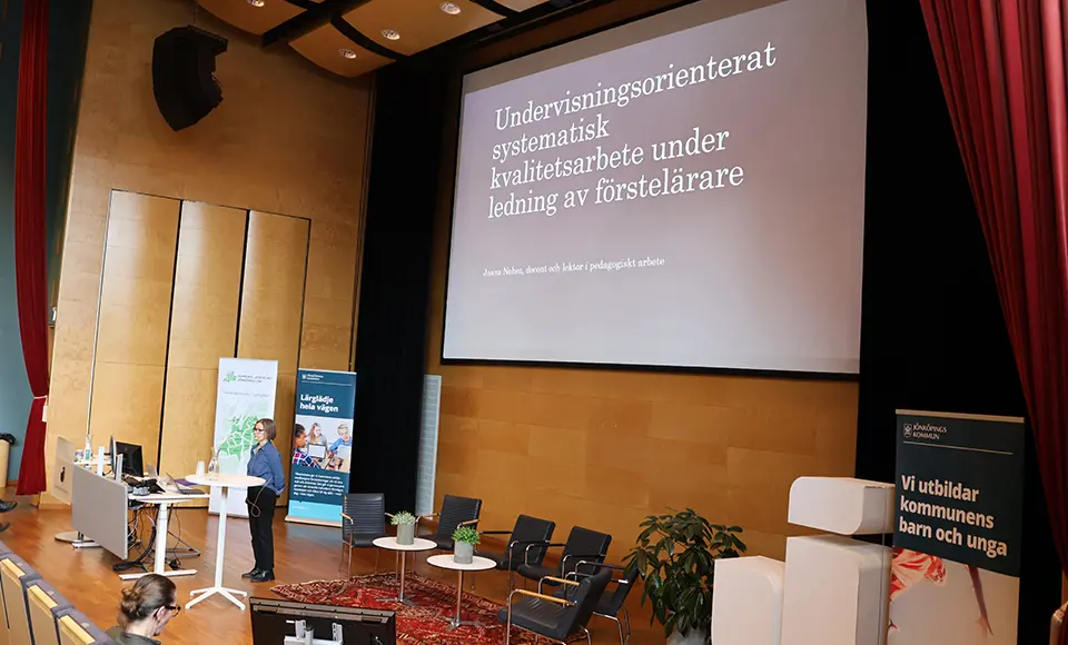 En person står på en scen och håller en föreläsning. På väggen står Undervisningsorienterat systematiskt kvalitetsarbete under ledning av förstelärare