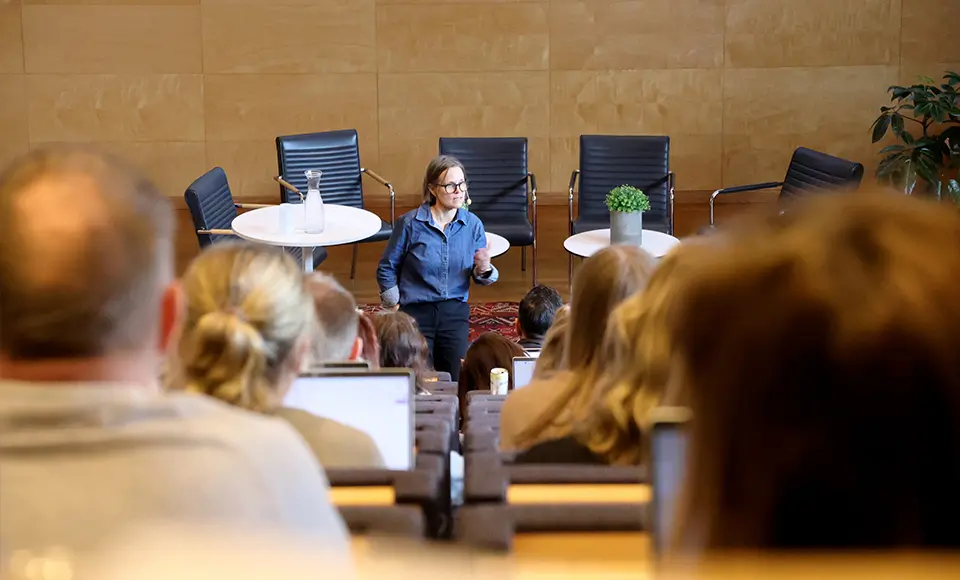 Ryggar på personer som sitter i en aula. På scen står en person och pratar.