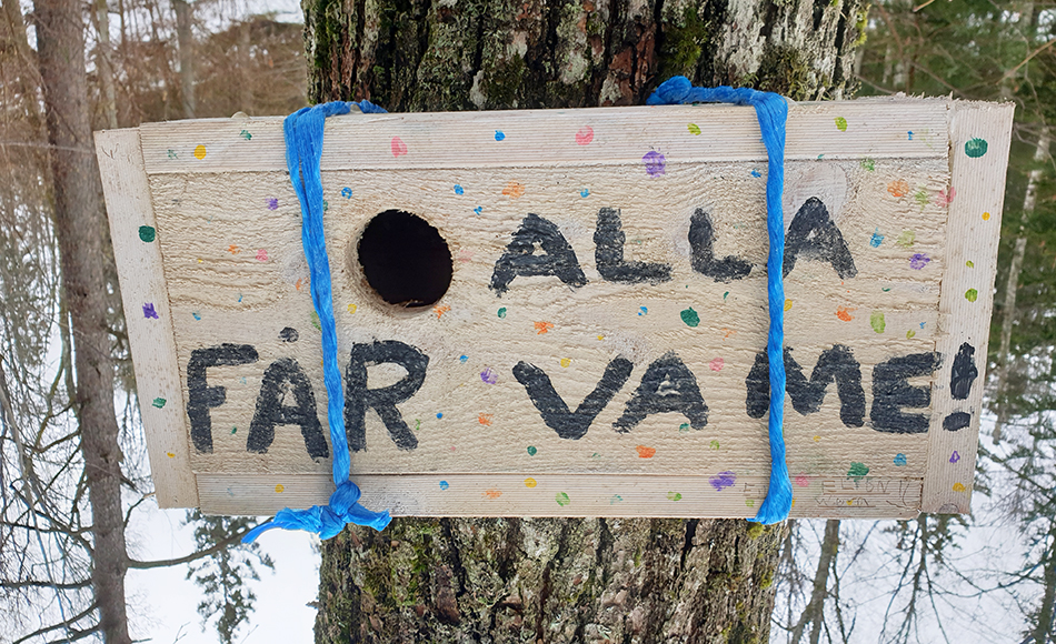 Foto på en skylt uppsatt på en trädstam. Texten lyder: alla får va me.