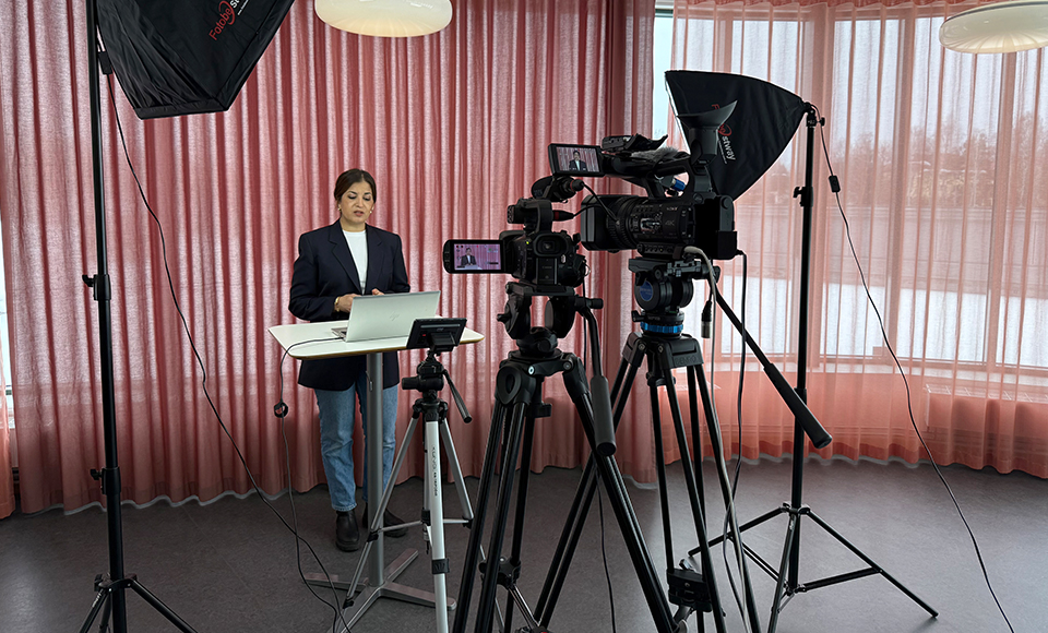 Filminspelning med två kameror på stativ och studiobelysning riktade mot en föreläsare som står vid ett bord med dator, framför rosa gardiner.