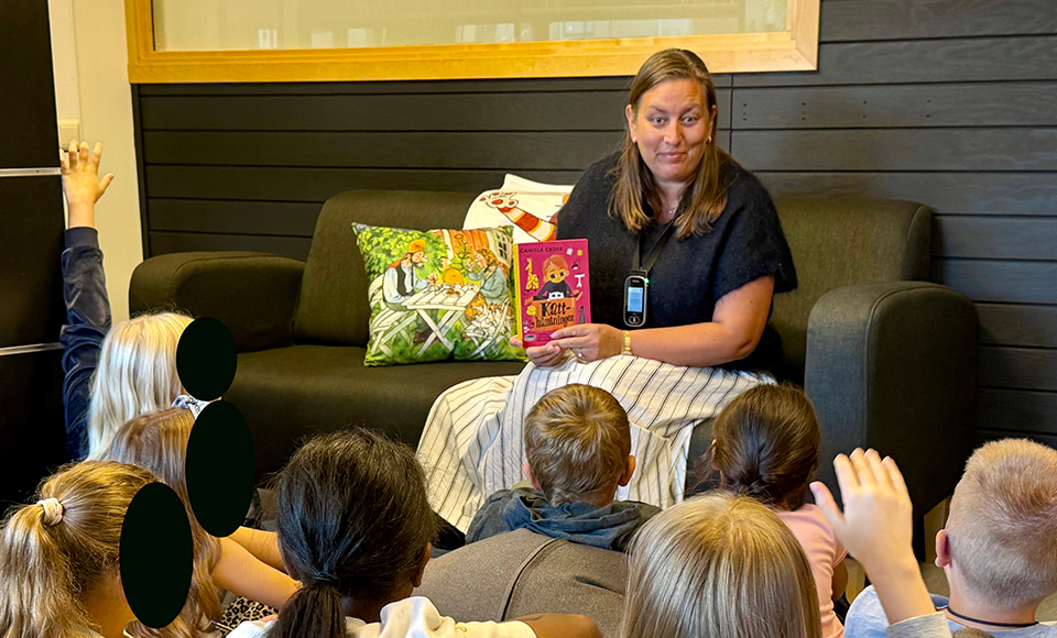 Johanna håller upp en bok under en högläsningsstund i skolbiblioteket. Flera barn sitter framför och en räcker upp handen.