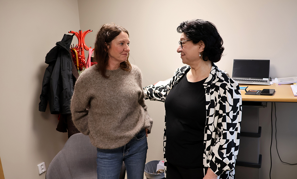 Charlotte och Amale står i ett kontorsrum. I rummet syns en stol, ett skrivbord med en laptop och papper, samt ytterkläder hängande på en krok i bakgrunden