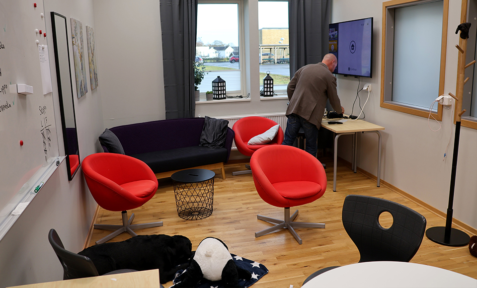 Ett rum med soffa, fåtöljer och en whiteboard. På väggen hänger en TV och Fredrik står bredvid och kopplar in Nintendospel.