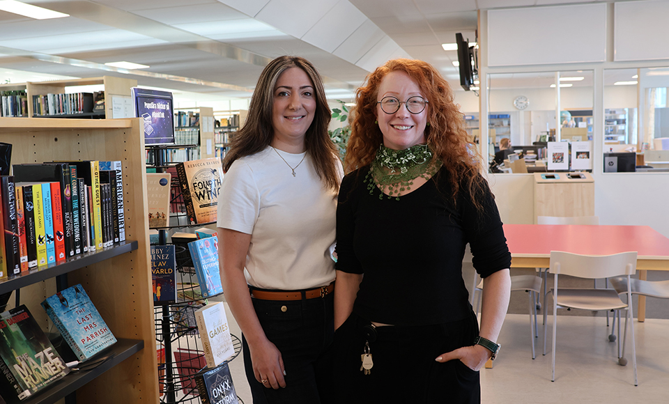 Två personer står bredvid varandra i ett skolbibliotek. I bakgrunden syns bokhyllor, bord och stolar i en öppen och ljus biblioteks­miljö.