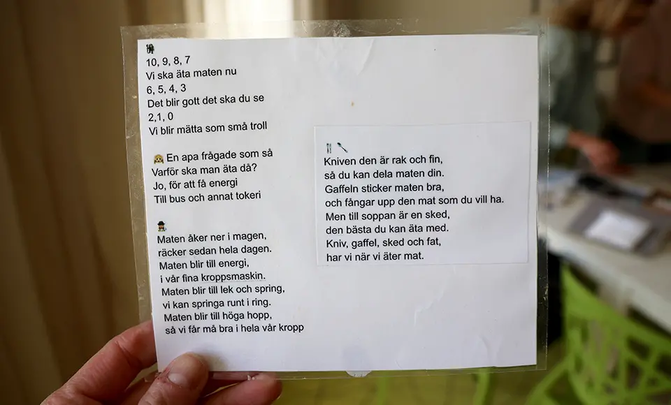 En hand håller upp ett laminerat papper med matramsor på svenska, dekorerat med små emojis. På vänstra sidan finns ramsor om varför vi äter mat och hur den ger energi, medan högra sidan har en dikt om kniv, gaffel och sked. Texten är tryckt i svart på vitt papper, och i bakgrunden syns suddigt ett rum med människor och gröna stolar.