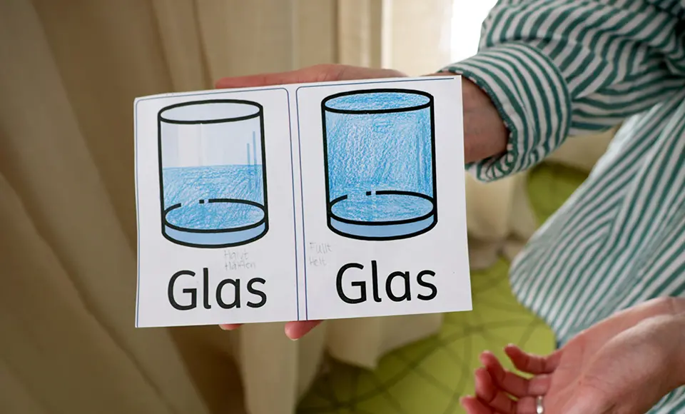 Två tecknade glas med vatten hålls upp av en person i randig tröja. Det vänstra glaset är halvfullt och märkt "Halv Full", medan det högra är helt fullt och märkt "Fullt Hel". Båda har texten "Glas" under sig