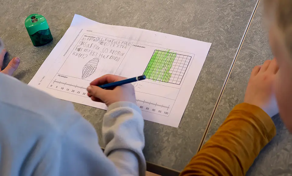 Man ser ryggen på två barn som sitter bredvid varandra. Framför dem ligger ett papper med en matematikuppgift. Den ena eleven håller i en penna.